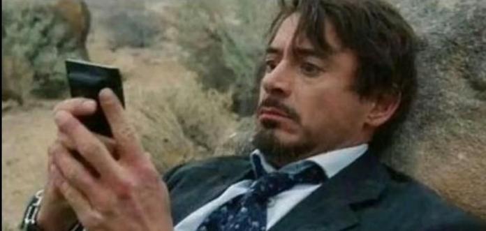 Un homme en costard est allongé au sol, adossé à une pierre, et regarde son téléphone qu'il tient dans ses mains. Il a les yeux légèrement écarquillés et une légère expression de peur sur le visage