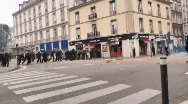 Vue de côté. À gauche une ligne de CRS avance en courant sur une personne en noir, face à eux, statique, bras tendus en croix, à droite de l'image. La ligne la bouscule puis se stoppe. 
Travelling avant. Les CRS reculent. Il avance dans leur direction. Travelling latéral qui tourne autour de la scène pour se placer derrière. Une personne masquée mais habillée de brun avance et se place bras-dessus bras-dessous avec le manifestant en noir.  Ils sont au centre de l'image, face aux keufs. Ils avancent.
Dézoom et travelling avant pour les suivre. À droite de l'image une autre personne arrive et donne un coup de pied dans un palet de lacrymo qui attérissait à peine aux pieds des deux premiers.
À gauche de l'image un manifestant cagoulé portant un gilet jaune rejoint bras dessus bras dessous les deux autres. On voit une autre personne approcher pour les rejoindre.