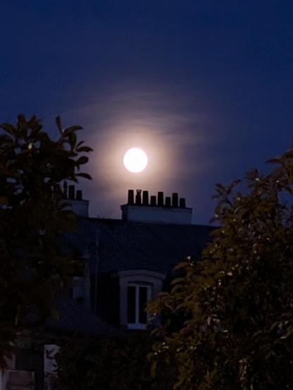 une lune pleine et ronde sous un voile nuageux, au dessus de cheminées d'appartements, sous un ciel bleu nuit